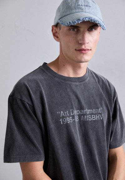 Junger Mann trägt ein ausgewaschenes schwarzes T-Shirt "Art Department 1995-08 MISBHV" und eine hellblaue, ausgefranste Jeansmütze vor einem schlichten grauen Hintergrund.