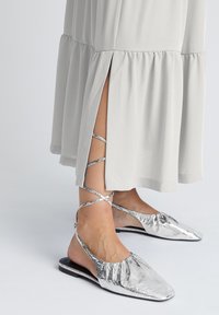 Ballerines argentées à finition texturée, dos ouvert, avec des brides croisées à la cheville, associées à une robe en tissu gris clair plissé.
