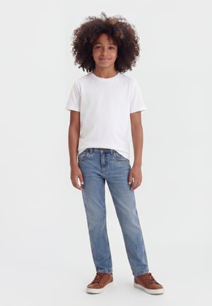 Παιδί με σγουρά μαλλιά που φοράει άσπρο μπλουζάκι, μπλε τζιν και καφέ αθλητικά παπούτσια, στέκεται μπροστά σε ένα απλό λευκό φόντο.