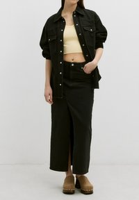 Zwarte denimjack over een gele geribbelde crop top, gecombineerd met een zwarte denimrok met een zijsplit en bruine klompen met studs.