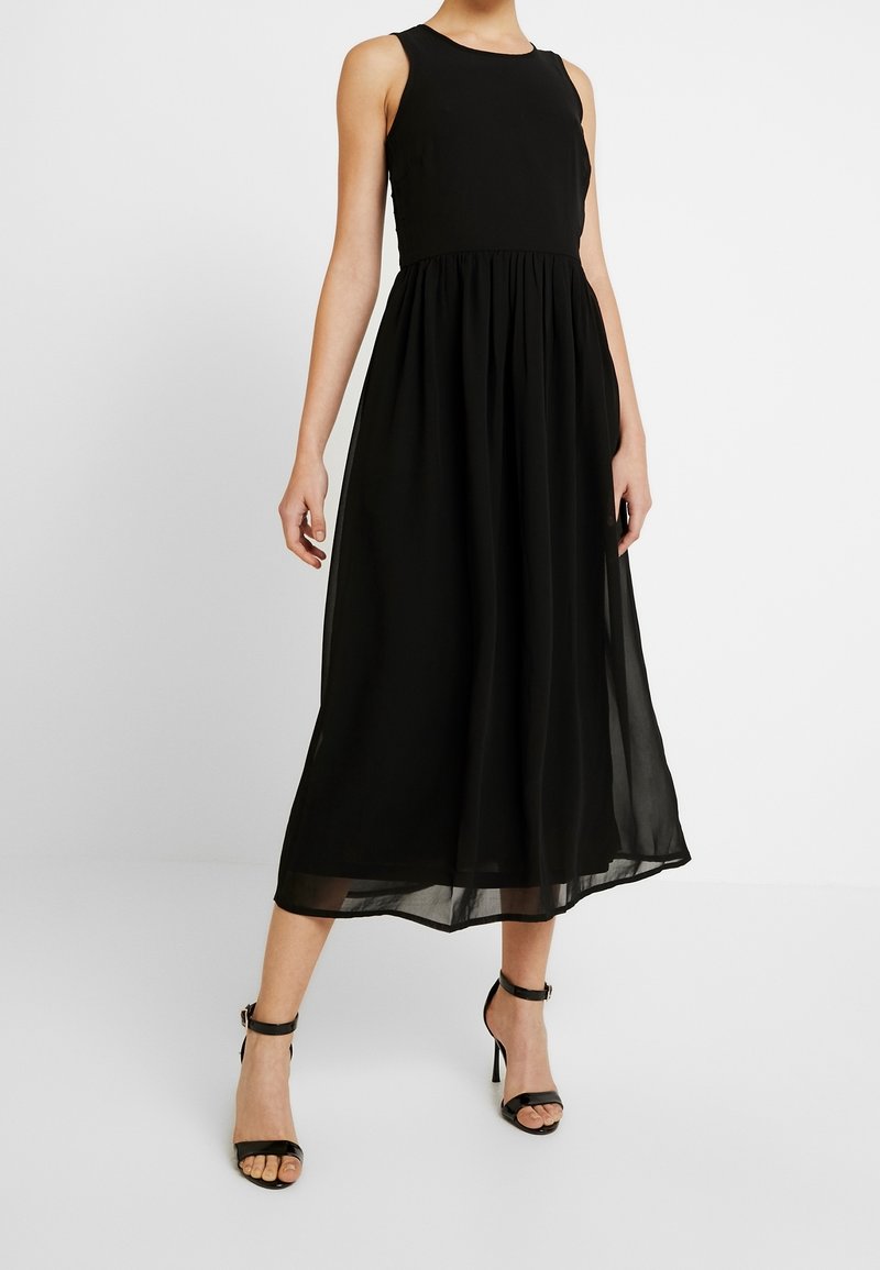 Femme portant une robe midi noire sans manches avec taille froncée et des sandales à talons hauts et brides à la cheville, sur fond blanc.