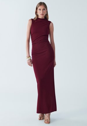 Burgundy sleeveless maxi dress with a fitted silhouette, smooth texture, and draped detail at the waist. Paired with strappy neutral heels.