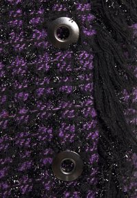 Gros plan d'un tissu noir et violet texturé avec deux boutons noirs brillants et des pompons en fil noir sur le côté droit.