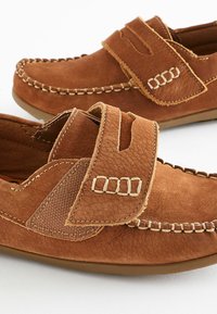 Chaussures à enfiler en cuir marron avec une surface texturée, des accents cousus et une sangle Velcro. Elles présentent une pointe ronde et une semelle flexible.