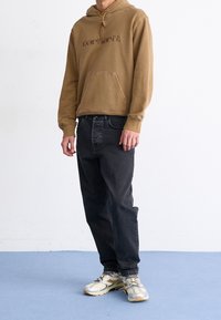 Henkilö, jolla on päällään ruskea Carhartt-huppari, mustat farkut ja valko-harmaat tennarit, seisoo vaaleansinisellä lattialla valkoista seinää vasten.