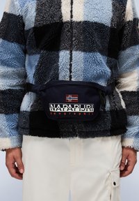 Marsupio blu navy con logo "NAPAPIJRI geographic" e patch con la bandiera norvegese, indossato sopra una giacca in pile a scacchi neri e blu.
