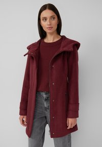 Burgunderrot melierter Wollmantel mit Kapuze, Knopfverschluss und seitlichen Reißverschlusstaschen; getragen über einem passenden Pullover und grauen Jeans.