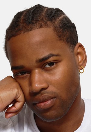 Portrait of a person with short, braided black hair and a gold hoop earring, resting their chin on a light-colored shirt-clad arm.