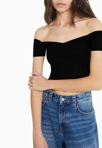 Femme portant un crop top noir court à épaules dénudées et un jean taille haute bleu, se tenant avec un bras croisé sur l'autre.