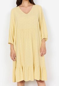 Robe jaune à col en V avec un motif géométrique, manches 3/4, taille élastiquée et ourlet volanté ; fabriquée en tissu léger.