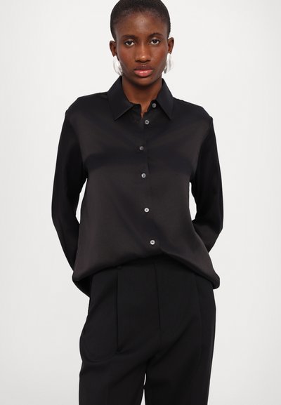 Chemise noire à manches longues et boutons en tissu lisse, dotée d'un col et d'un léger éclat, associée à un pantalon noir taille haute.