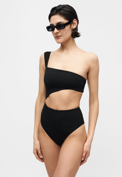 Γυναίκα με μαύρο ολόσωμο μαγιό με άνοιγμα σε έναν ώμο και μαύρα ορθογώνια γυαλιά ηλίου στέκεται μπροστά σε απλό φόντο.