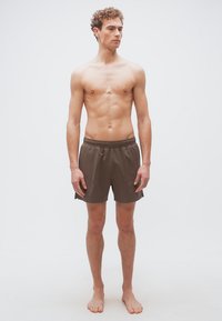 Νεαρός άνδρας όρθιος, ξυπόλυτος, φοράει καφέ σορτς με ελαστική μέση, κοιτάζοντας ελαφρώς προς το πλευρό σε απλό λευκό φόντο.