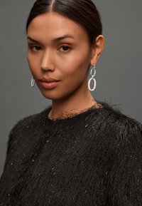 Prenda negra con textura y detalles de flecos, combinada con pendientes de aro plateados que presentan un patrón decorativo. Fondo neutro.