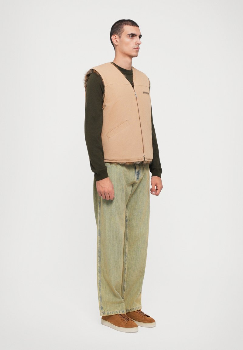 Beige sleeveless vest with side pockets and a smooth texture, paired with light green striped pants and brown sneakers.