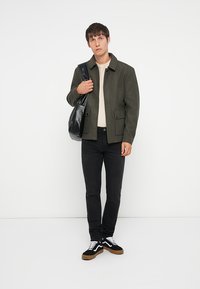 Olijfgroene wolmix jacket met een kraag, twee frontritsen, crèmekleurig shirt met ronde hals, zwarte slim fit jeans en zwarte sneakers met witte accenten.