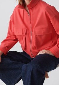 Personne portant une veste zippée rouge vif avec de grandes poches frontales et un jean large bleu foncé, assise devant un fond uni.