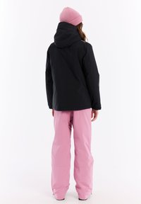 Zwarte waterdichte jas met een capuchon, gecombineerd met twee roze sneeuwbroeken. De jas heeft elastische manchetten en de muts is ook roze en gebreid.