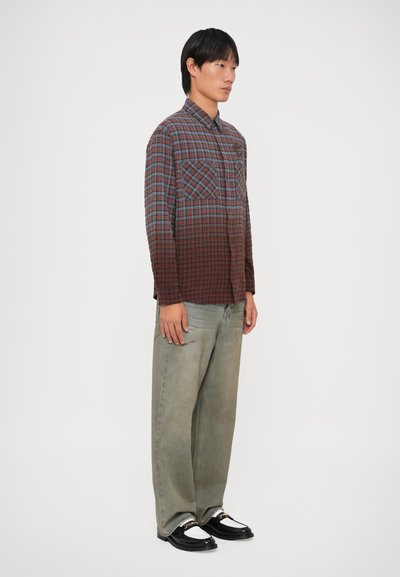 Geruite shirt met blauwe, rode en bruine tinten, lange mouwen, twee borstzakken; gecombineerd met gestructureerde lichtgroene spijkerbroek en zwarte schoenen.