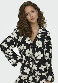 Schwarze Bluse mit weißem Blumenmuster, mit V-Ausschnitt, langen Ärmeln und Bindebändern vorne. Strukturierter Stoff mit lockerer Passform.