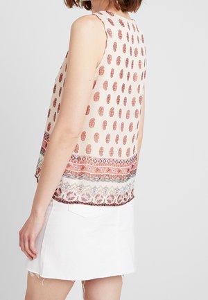 Femme portant un haut sans manches crème avec un imprimé bordure cachemire et floral rouge, associé à une jupe blanche sur un fond uni.