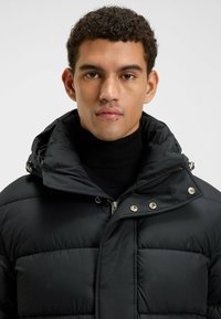 Chaqueta acolchada negra con cuello alto, que presenta un cierre de cremallera y acentos en dorado. Diseño texturizado y acolchado con un acabado suave.