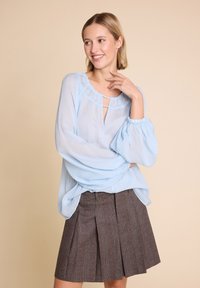 Kvinne som smiler, iført en lyseblå gjennomsiktig bluse med puffede ermer og et brunt plissert skjørt, stående mot en beige bakgrunn.