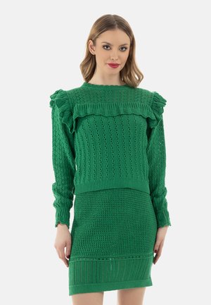 Vrouw draagt een groene gebreide trui met ruches op de schouders en een bijpassende getextureerde gebreide rok.