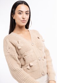 Béžový pletený cardigan s V výstřihem, se strukturovanými vzory a mašlovými akcenty. Má velké knoflíky s perlovými detaily.