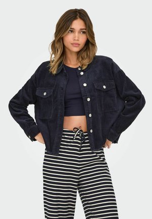 Navy corduroy jacket with two chest pockets and silver snap buttons; paired with black and white striped relaxed-fit trousers.