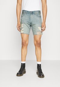 Person wearing light blue distressed denim shorts, white socks, black lace-up boots, and a white shirt, standing against a plain background.