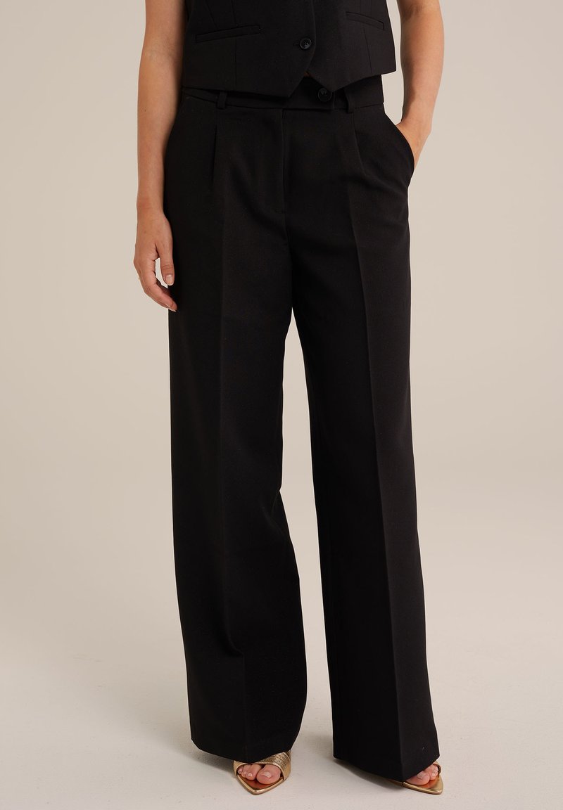 Črne široke hlače z gladko teksturo, ki imajo stranske žepke in prilagojen kroj, dopolnjene z oprijetim jopičem in sandali z odprto konico.