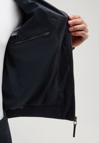 Marineblau bomberjacke mit einem glatten Außenmaterial. Das Innere verfügt über eine mit Reißverschluss gesicherte Tasche und ein strukturiertes Futter mit einem subtilen Muster.