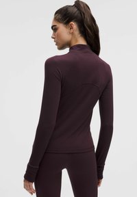 Ilga piedurknju sporta krekls tumši burgundijas krāsā, izgatavots no gludas stiepjamās auduma, ar augstu apkakli un plecu šuvju detaļām.