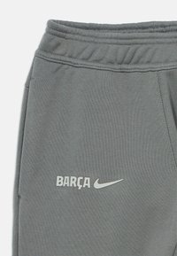 Sivé bavlnené tepláky s elastickým pásom. Na dolnej ľavej strane predného dielu sú potlačené "BARÇA" a logo Nike v bielej farbe. Hladká textúra.
