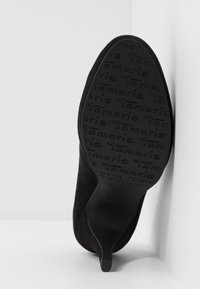 Chaussure à talon haut en velours noir inclinée contre un mur blanc, semelle montrant un motif de marque "Tamaris" texturé et taille 37.