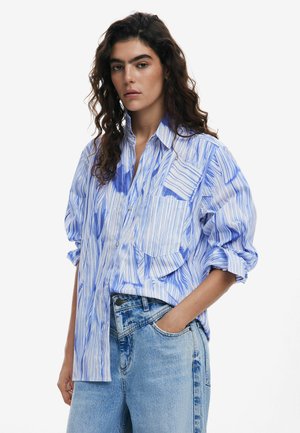 Blue and white striped oversized shirt with a button-down collar, featuring large patch pockets and rolled-up sleeves, paired with denim jeans.