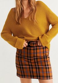 Frau in senffarbenem, geripptem Pullover und einem hochtaillierten Rock aus orangefarbenem, schwarzem und blauen Karomuster mit einem schwarzen Gürtel.