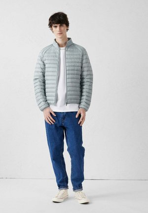 Jonge man staand, gekleed in een lichtgrijs gewatteerd jack, wit T-shirt, blauwe spijkerbroek en witte sneakers tegen een effen witte achtergrond.