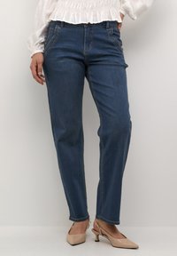 Jeans in denim blu con taglio dritto e dettagli di cucitura visibili. Indossati con una blusa bianca con rouches e scarpe con tacco color nude.