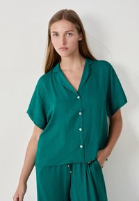Jonge vrouw met lang blond haar, gekleed in een teal korte mouwen overhemd met knopen en bijpassende losse broek, staand tegen een effen muur.