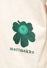 Zöld absztrakt virág kör alakú központtal, a "marimekko" felirat fölött, világos bézs színű vászon háttéren.