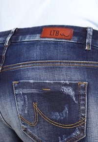 Vaqueros de mezclilla en lavado oscuro con desgastado. Presentan un parche de cuero marrón etiquetado "LTB" y costuras naranjas en el bolsillo trasero con un diseño curvo.