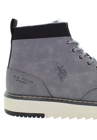 Šedá kotníková obuv U.S. Polo Assn. s černým vroubkovaným límcem, bílou podešví, kovovými očky a logem hráče polo na boku.