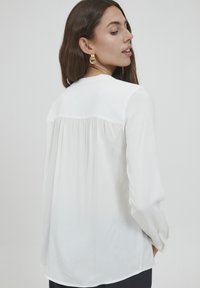 Witte blouse met lange mouwen en een geplooid rugdetail, zachte textuur, afgeronde zoom en knoopsluiting bij de manchetten. Model wordt van achteren getoond.