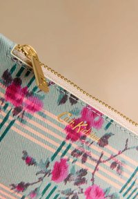 Gėlių raštų užtrauktuku įsegta dėklas šviesiai žalsvos spalvos, puoštas rožinėmis rožėmis ir žaliomis juostelėmis. Auksinės spalvos užtrauktukas ir audinio logotipo detalė.