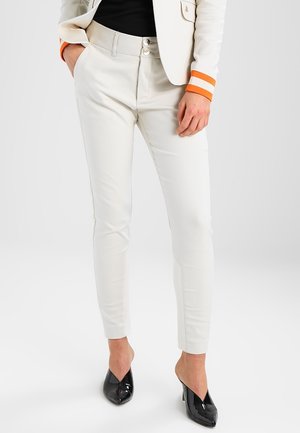 Kvinde iført tætsiddende hvide bukser med dobbelte knapper i taljen, cremefarvet blazer med orange manchetter og sorte spidse høje hæle.