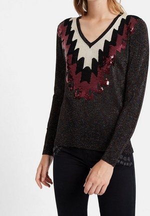 Femme portant un pull noir à col en V avec des motifs zigzag à paillettes en rouge, noir et blanc, assorti à un pantalon noir.