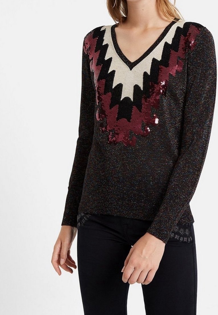 Femme portant un pull noir à col en V avec des motifs zigzag à paillettes en rouge, noir et blanc, assorti à un pantalon noir.