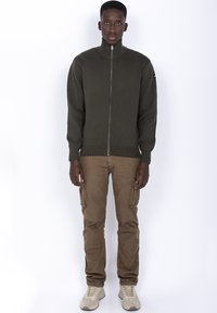 Mørkegrønt zip-up sweater med høj krave, båret over beige cargo bukser og beige sneakers. Stoffet ser glat og let struktureret ud.
